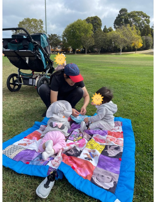 A caregiver sitting with young children on a blanket in the park; the children’s faces have stickers