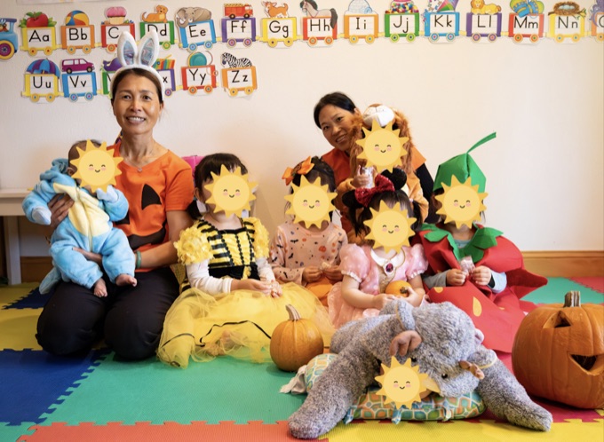 Caregivers and children in costumes in the daycare playroom; the children’s faces have stickers