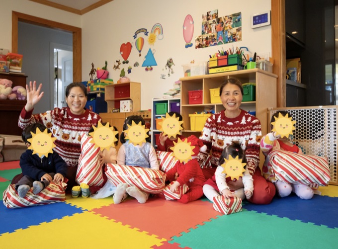 Children and caregivers in festive outfits sitting on the playroom mat; the children’s faces have stickers